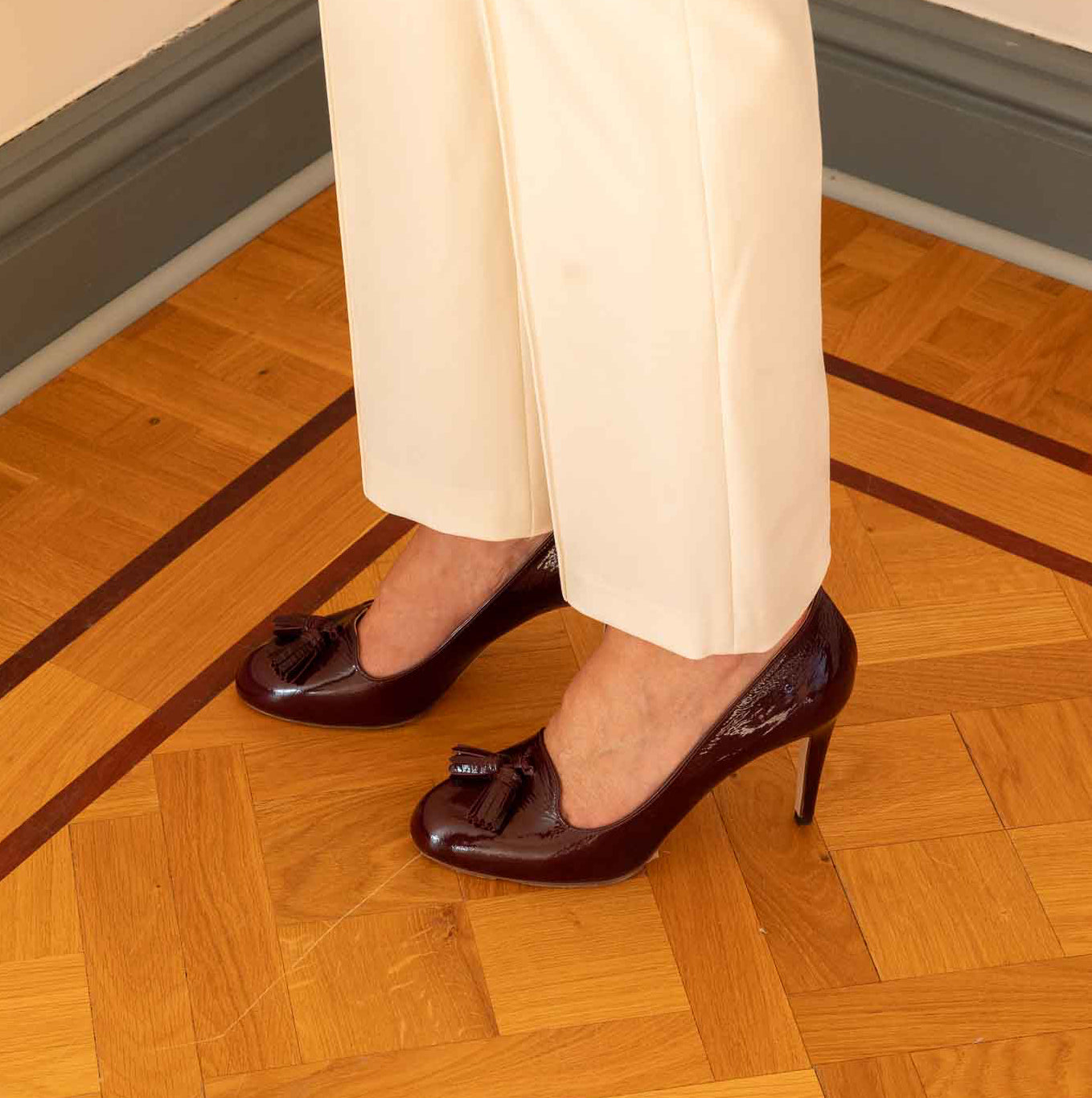 A woman wearing dark burgundy patent leather kitten-heel pumps with tassel details, paired with cream-colored trousers, standing on a polished wooden parquet floor.