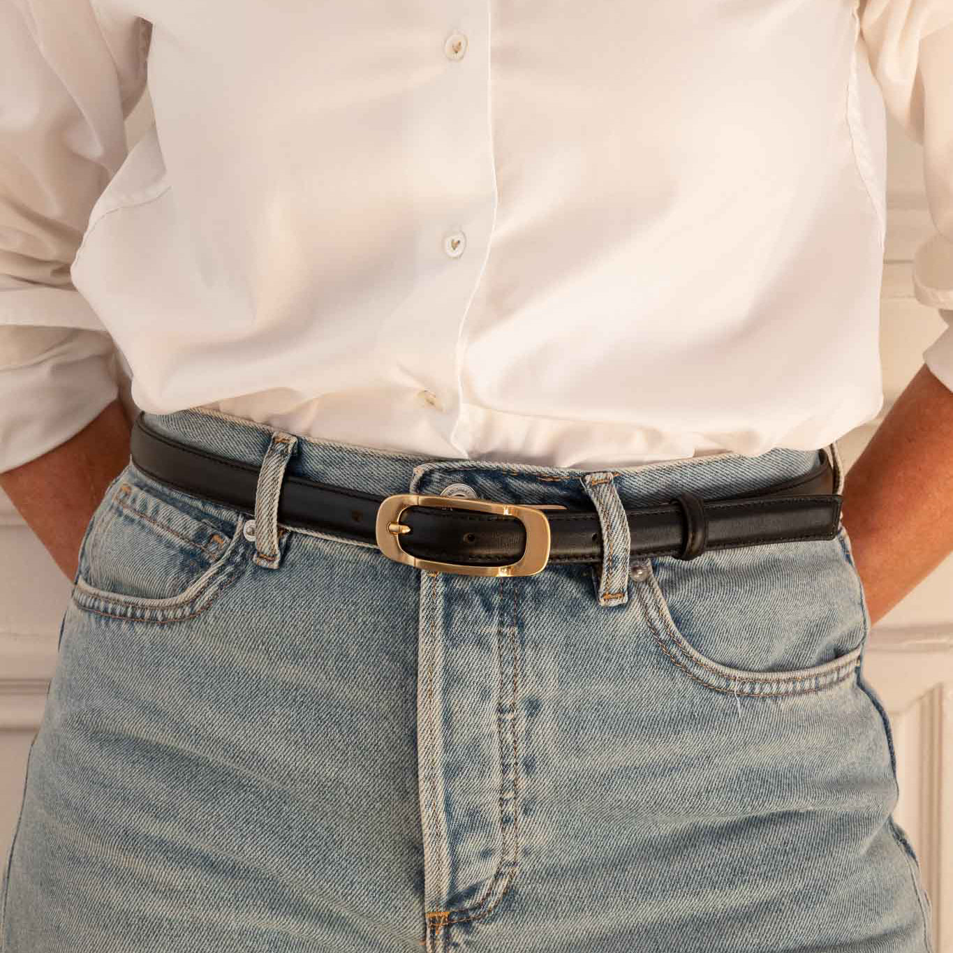 A close-up of a person wearing a black leather belt with a gold-tone rectangular buckle, tucked into light-wash blue jeans and paired with a white button-down shirt.