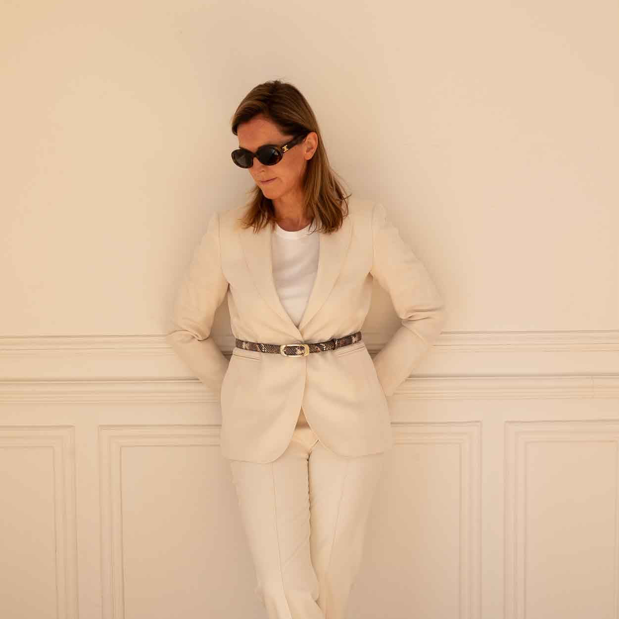A woman wearing a cream-colored two-piece suit with a white top, a winter python printed belt, and patterned heels, posing against a cream paneled wall on a wooden parquet floor.