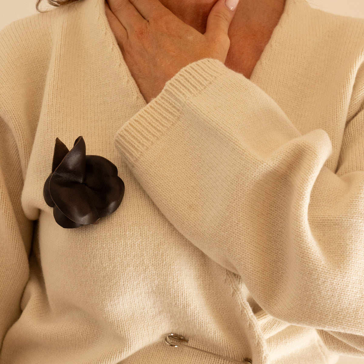 Close-up of a cream knit cardigan worn by a model, featuring a dark brown nappa leather rose brooch pinned to the chest and a silver safety pin detail at the front.
