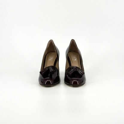 A pair of dark burgundy patent leather pumps with tassel details on the toes, shown from the front side against a plain white background.