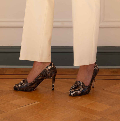 A person wearing cream-colored trousers and black and brown snakeskin-patterned high-heeled pumps, standing on a wooden parquet floor against a cream wall.