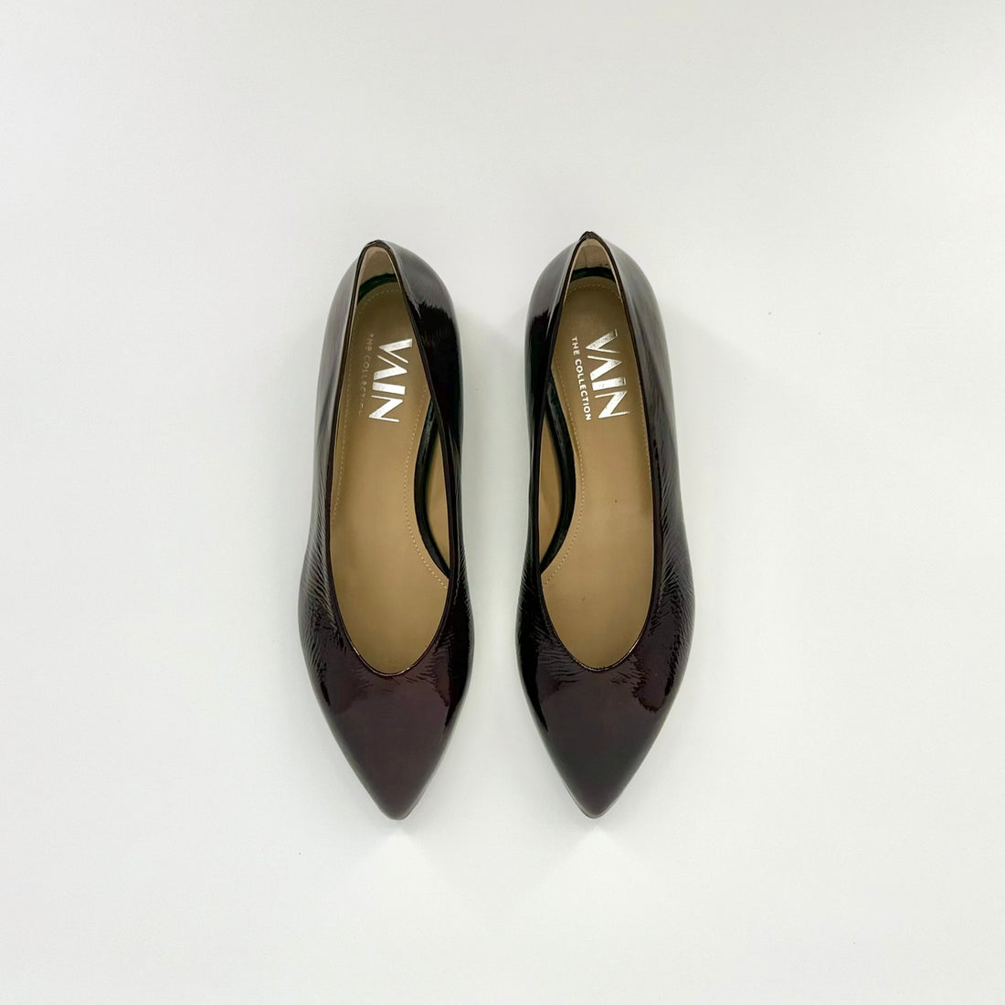 A pair of pointed-toe ballerinas in a glossy dark bordeaux/plum-brown patent leather, shown from a top-down view on a light gray background. The brand name &