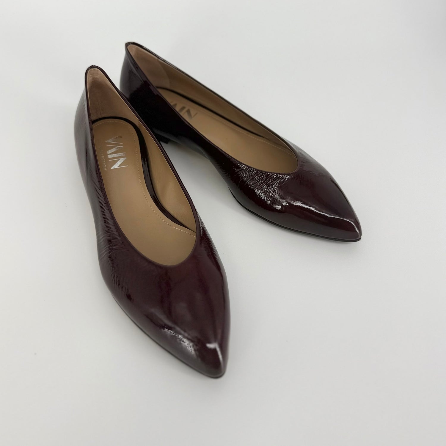 A pair of pointed-toe ballerinas in a glossy dark bordeaux/plum-brown patent leather, shown on a light gray background. The brand name &