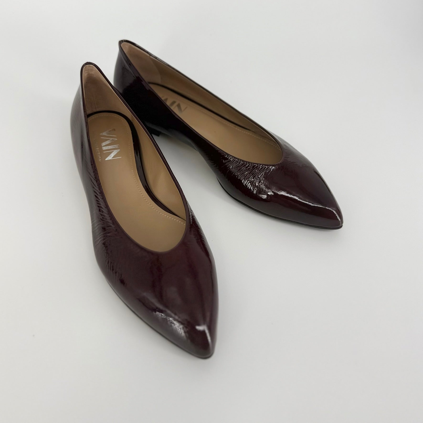 A pair of pointed-toe ballerinas in a glossy dark bordeaux/plum-brown patent leather, shown on a light gray background. The brand name &