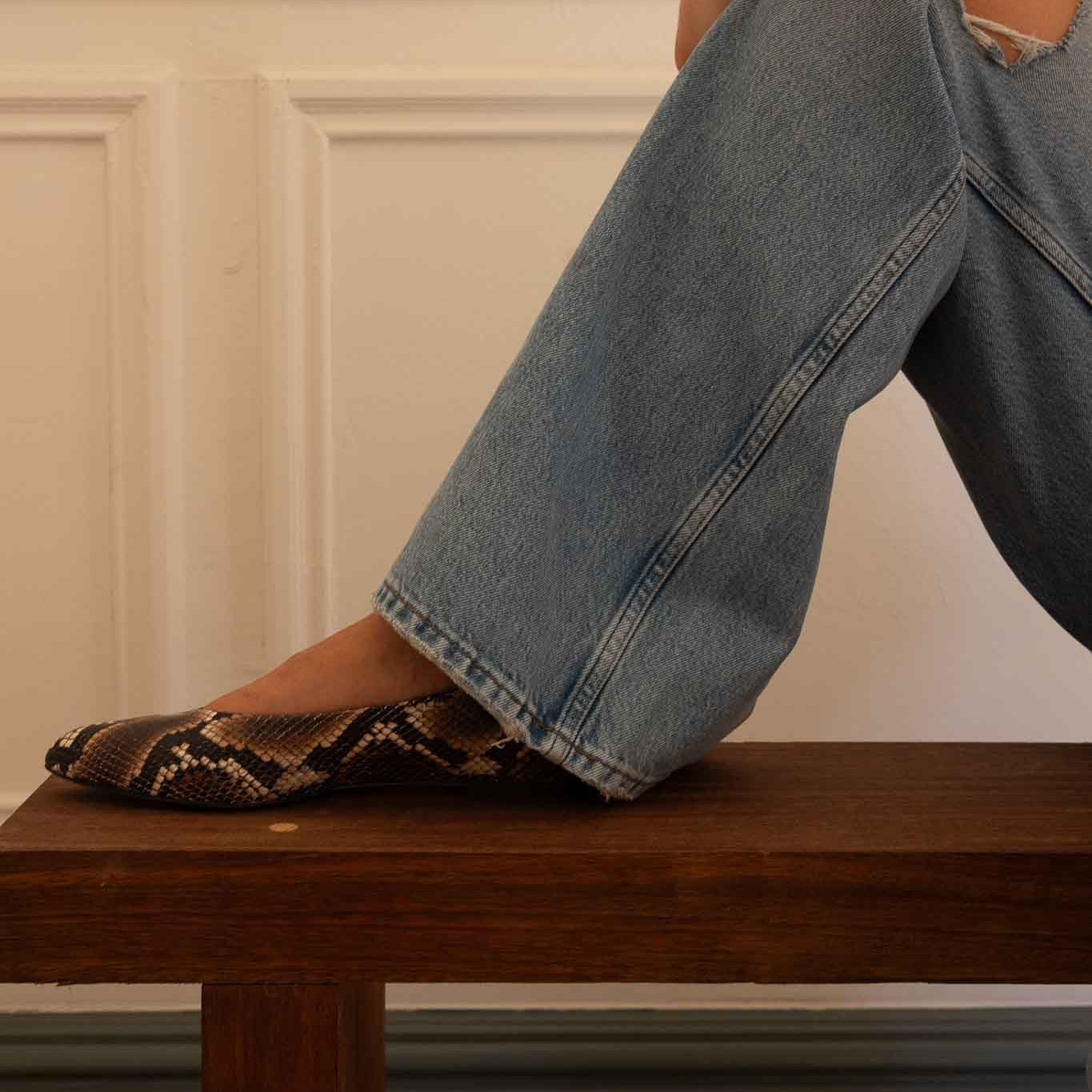 A woman sitting on a wooden bench wearing light-wash wide-leg jeans and a snakeskin-patterned flat shoe, posed against an off-white paneled wall.
