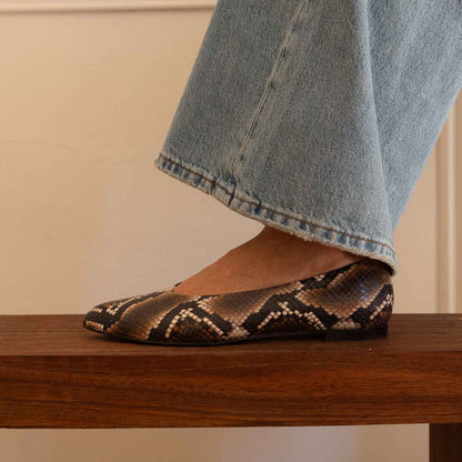 A woman wearing a snakeskin-patterned flat shoe with light-wash blue jeans, standing on a wooden bench against a cream-colored wall.