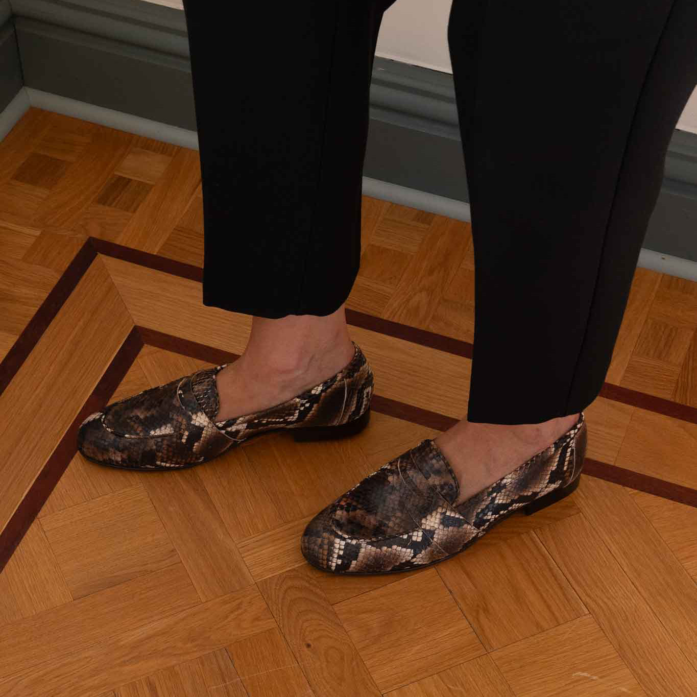 A woman wearing black trousers and snakeskin-patterned slip-on loafers, standing on a polished wooden parquet floor.
