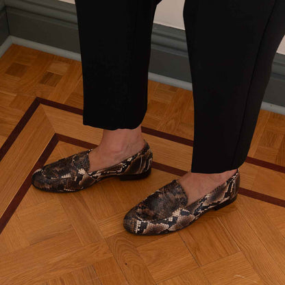 A woman wearing black trousers and snakeskin-patterned slip-on loafers, standing on a polished wooden parquet floor.