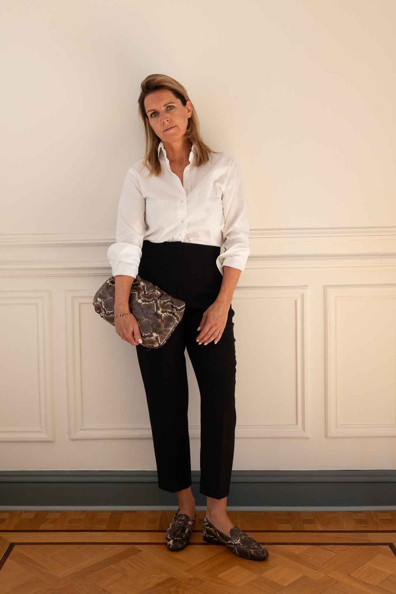 A woman stands indoors against a cream paneled wall, wearing a white button-up shirt, black cropped pants, and dark patterned loafers, while holding a brown and gray patterned clutch.