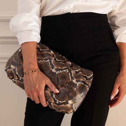 A person wearing a white button-up shirt and black trousers holds a snakeskin-patterned clutch bag against their hip.