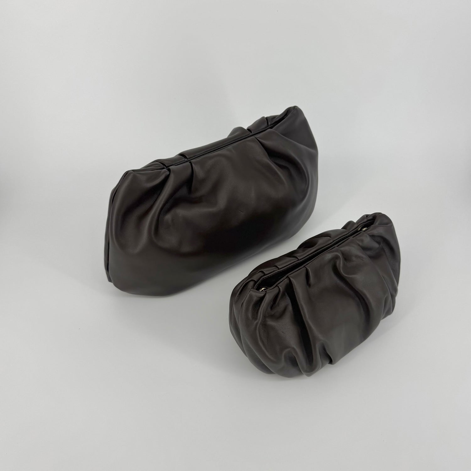 Two dark brown leather clutch bags, one small, the other larger, with a gathered, slouchy silhouette and magnetic closures, displayed on a light gray studio background.