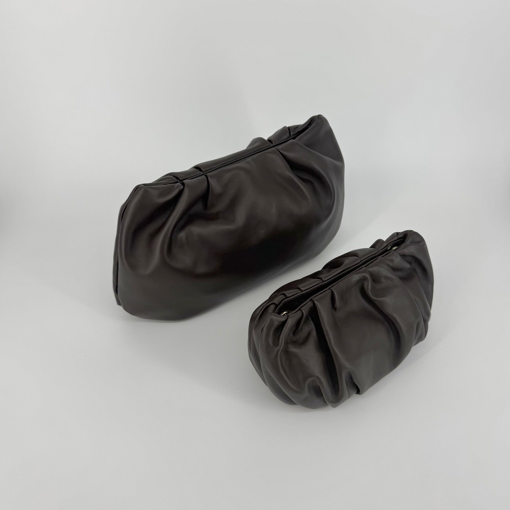 Two dark brown leather clutch bags, one small, the other larger, with a gathered, slouchy silhouette and magnetic closures, displayed on a light gray studio background.