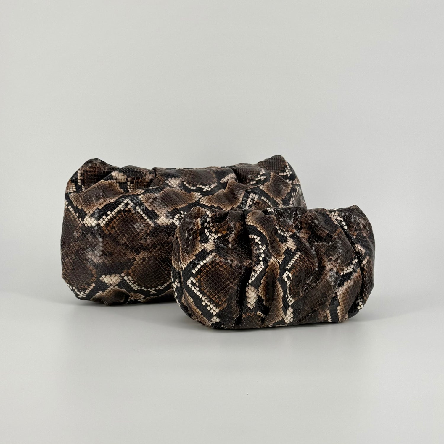 Mini and maxi snakeskin-patterned clutch bags in brown, black, and beige tones, photographed on a clean light gray studio background.