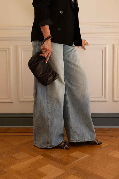Woman standing against a whit wall holding a darkbrown clutch wearing wide jeans and pointy kitten heels in warm winte python colours 