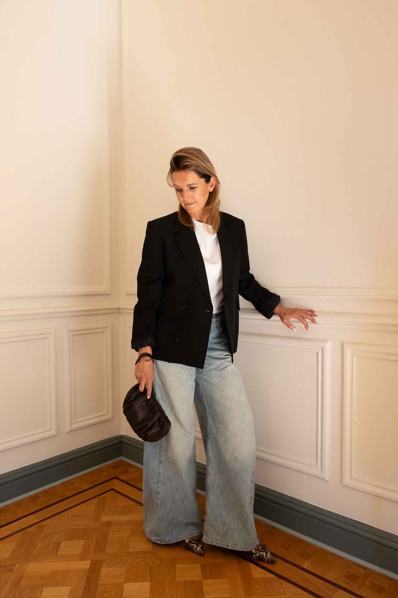 A woman stands in a corner of an indoor room with cream paneled walls and a wood parquet floor, wearing a black blazer over a white top, light-wash wide-leg jeans, and holding a dark brown handbag.