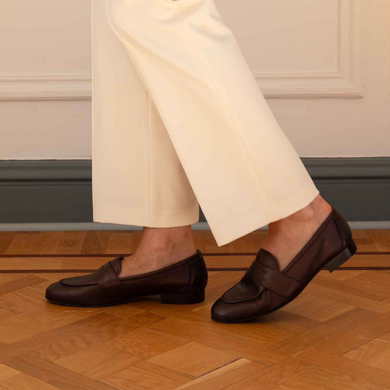 A person wearing cream wide-leg trousers and dark brown leather penny loafers, standing on a wooden parquet floor against a cream paneled wall.