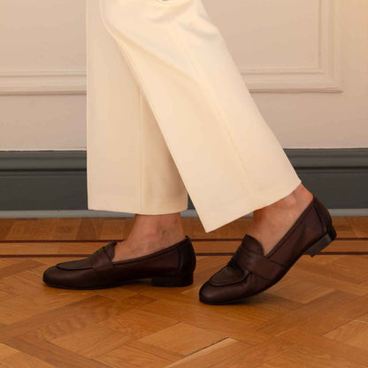 A person wearing cream wide-leg trousers and dark brown leather penny loafers, standing on a wooden parquet floor against a cream paneled wall.