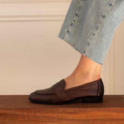 A person wearing light blue jeans and a dark brown leather penny loafer, standing on a wooden bench against a cream paneled wall.