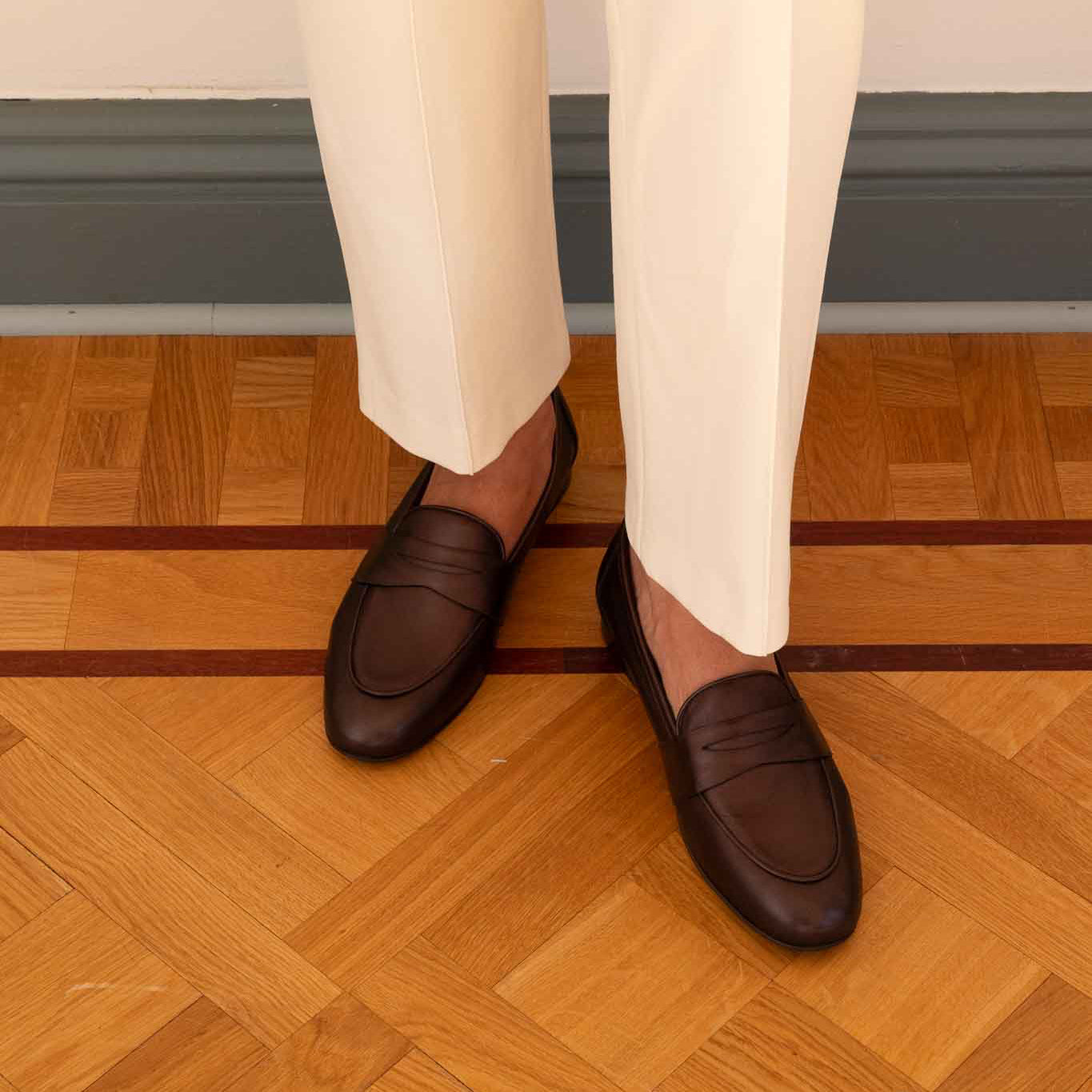 A person wearing dark brown leather penny loafers with cream trousers, standing on a polished wooden parquet floor.