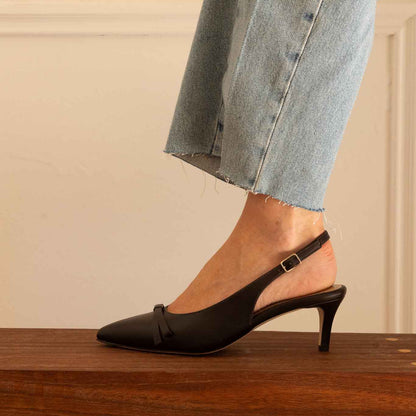A woman wearing light blue denim jeans and darkbrown slingback kitten heel pumps with a pointed toe, standing on a wooden bench against a cream-colored wall.