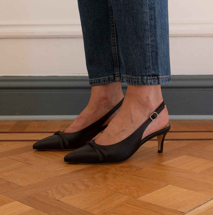 A woman wearing darkbrown slingback kitten heel pumps with ankle straps, paired with blue jeans, standing on a wooden parquet floor.