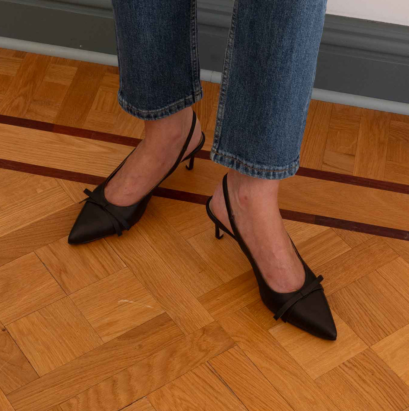 A woman wearing blue jeans and dark cacao brown pointed slingback heels with a small bow detail, standing on a wooden parquet floor.