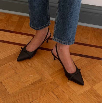A woman wearing blue jeans and dark cacao brown pointed slingback heels with a small bow detail, standing on a wooden parquet floor.