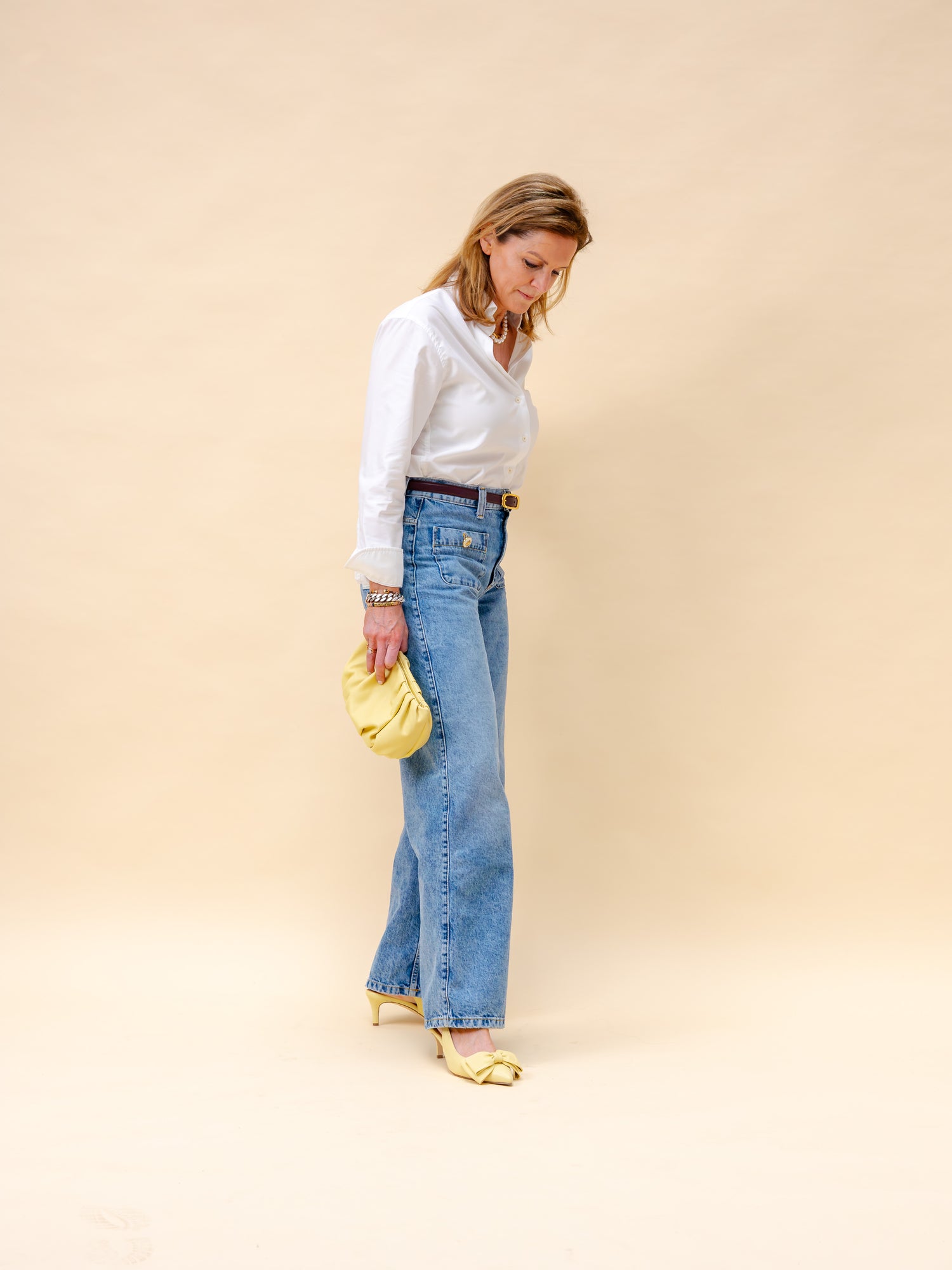 Woman wearing a white blouse and blue jeans and yellow low heels and matching clutch against a beige background