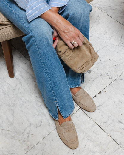 Woman wearing blue jeans and beige loafers sitting on a chair holding matching beige suede clutch.
