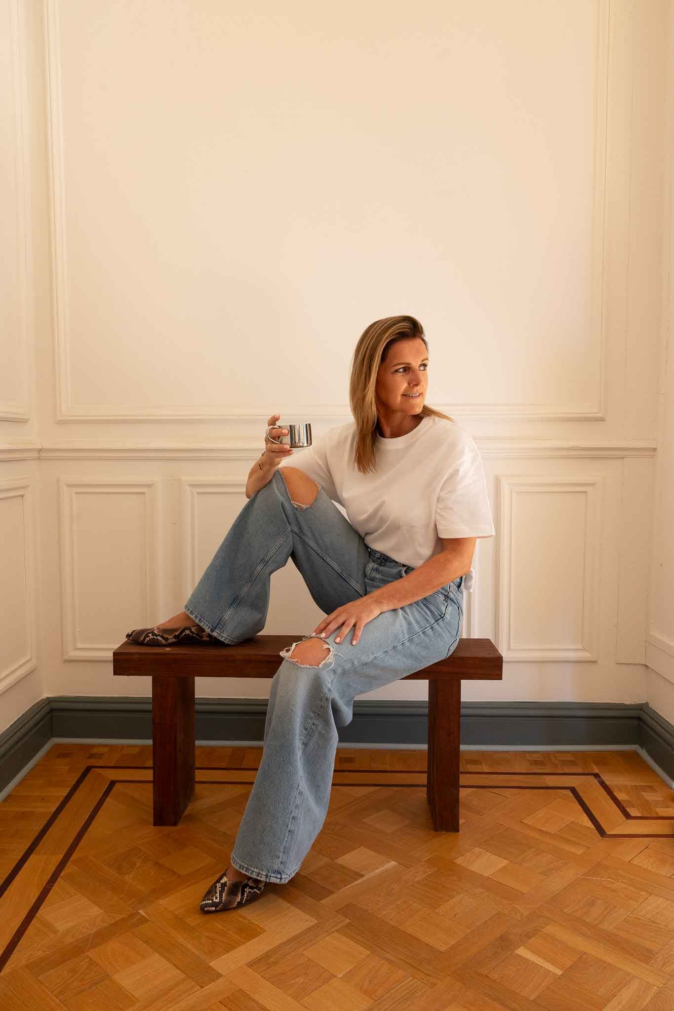 Woman sitting on a bench wearing lightblue jeans white t-shirt and winter python printed ballerinas drinking coffee from a stainless steel cup on a white background and parquet floor 