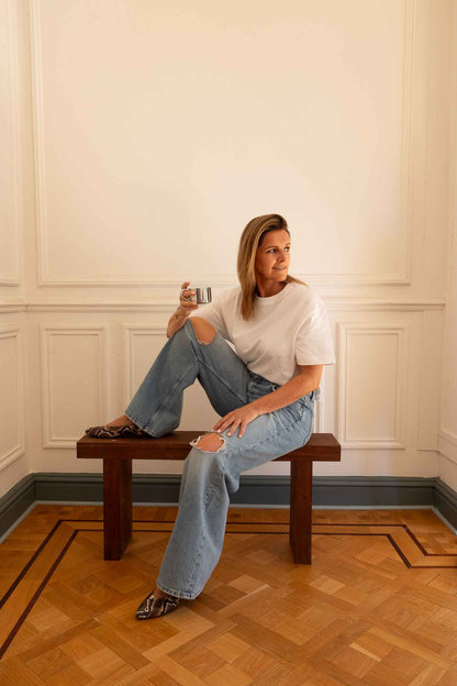 Woman sitting on a bench wearing lightblue jeans white t-shirt and winter python printed ballerinas drinking coffee from a stainless steel cup on a white background and parquet floor 