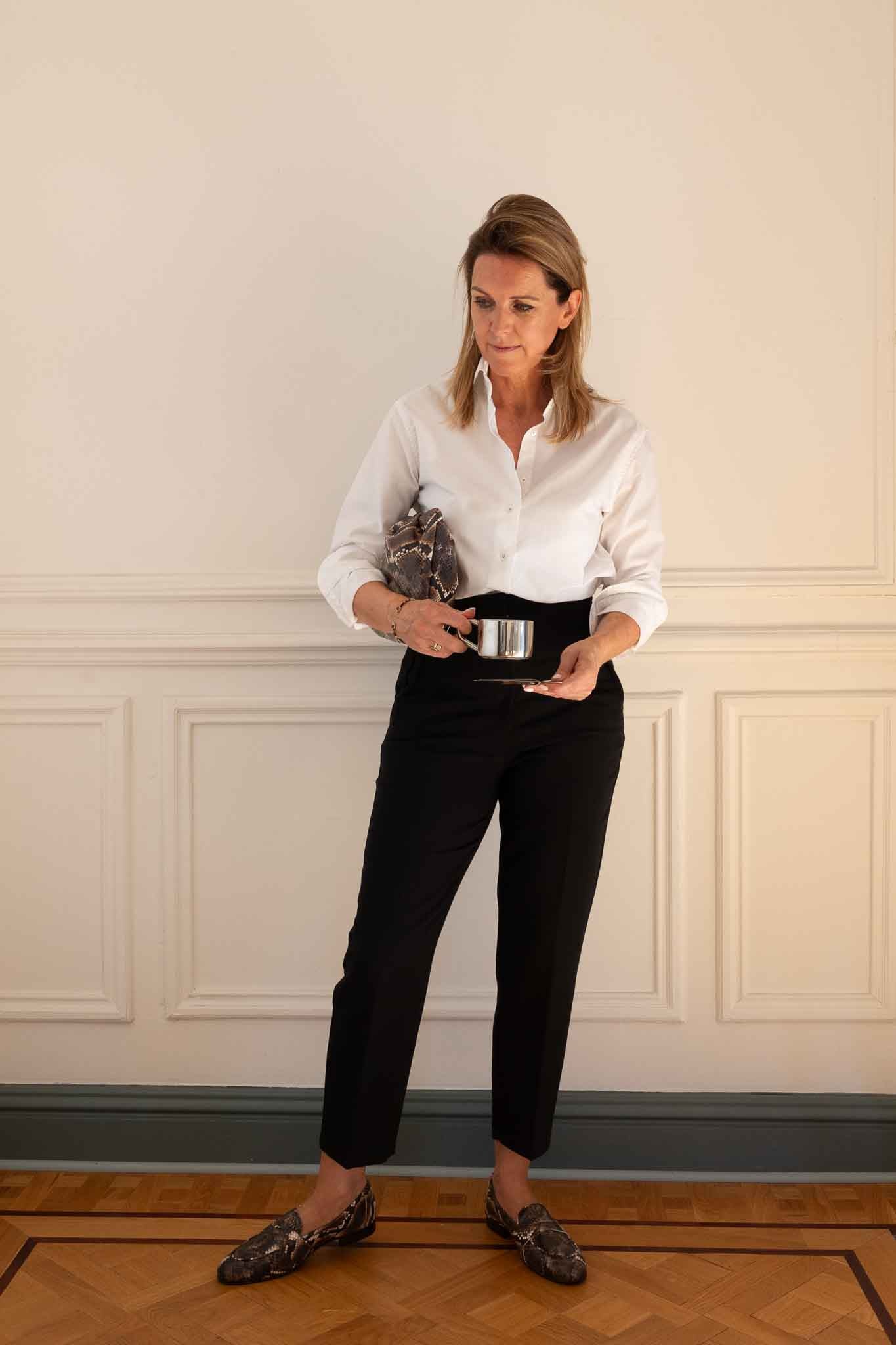 A woman stands in a room with cream paneled walls and a wood floor, wearing a white button-up shirt, black trousers, and dark patterned loafers while holding a small metallic object.